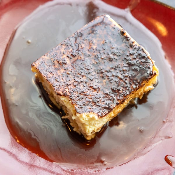 A square dessert with a caramelized top sits in a pool of dark chocolate sauce on a red plate.