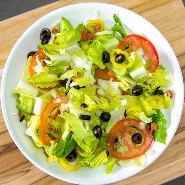 A fresh garden salad with lettuce, tomatoes, black olives, banana peppers, and cheese in a white bowl.