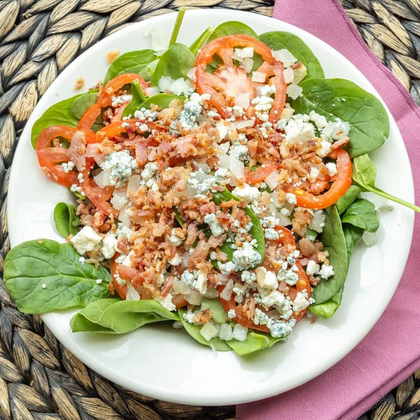 Spinach salad with sliced tomatoes, crumbled bacon, diced onions, and blue cheese on a white plate.