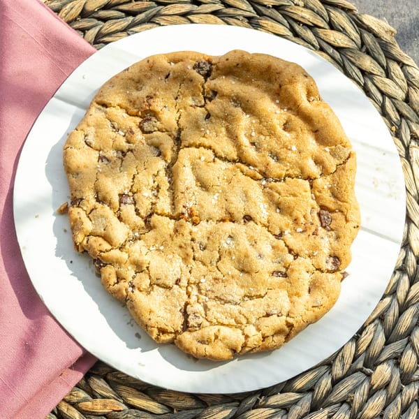 A large, golden-brown chocolate chip cookie with sea salt on a white plate.