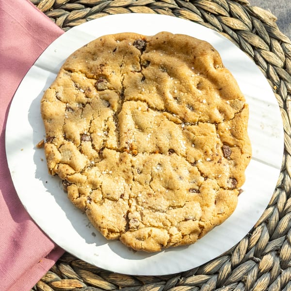 A large, golden-brown chocolate chip cookie with sea salt on a white plate and woven mat.