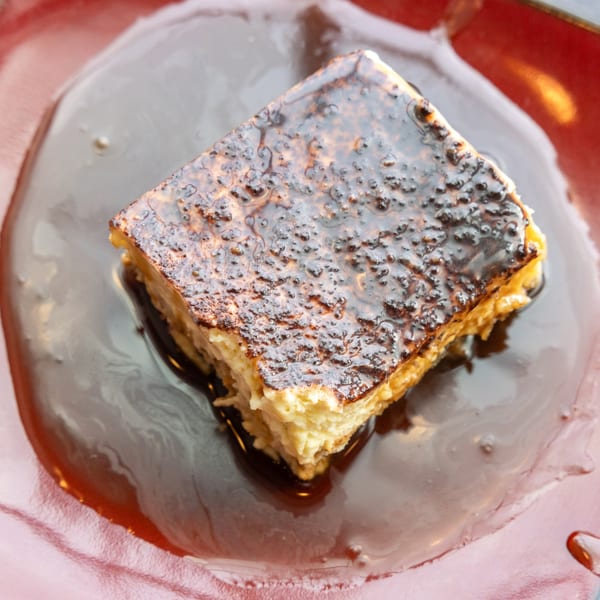 A square dessert with a caramelized top, sitting in a pool of dark sauce on a red plate.