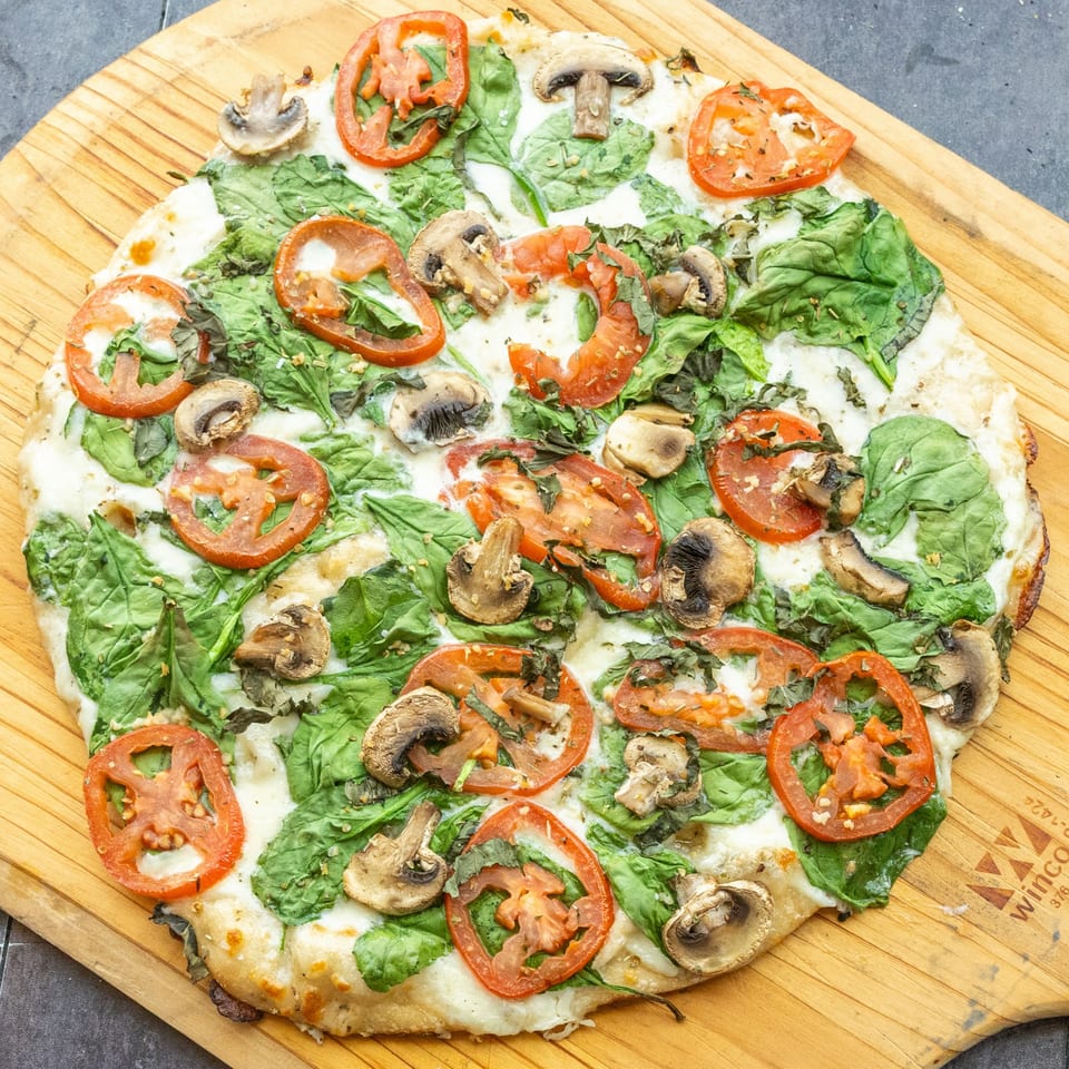 Mama Mimi's white pizza with spinach, sliced tomatoes, and mushrooms on a wooden peel.
