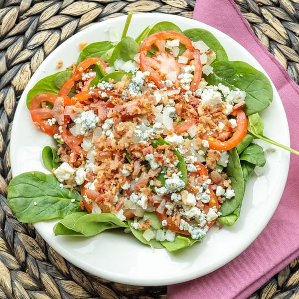 Spinach salad with sliced tomatoes, crumbled bacon, blue cheese, and diced onions on a white plate.
