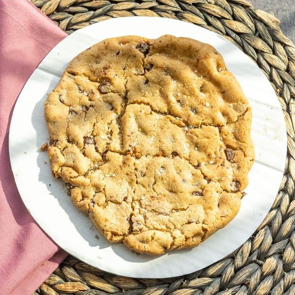 A large, golden-brown chocolate chip cookie with sea salt on a white plate and woven placemat.
