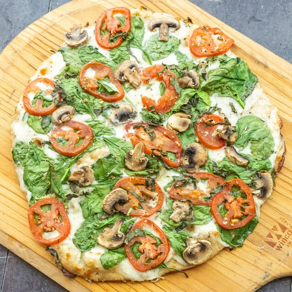 Mama Mimi's Take 'N Bake Pizza with spinach, mushrooms, and sliced tomatoes on a wooden board.