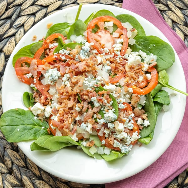 Spinach salad with sliced tomatoes, crumbled bacon, blue cheese, and diced onions on a white plate.