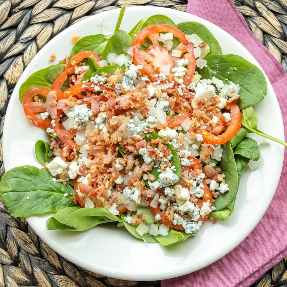 Spinach salad with sliced tomatoes, crumbled bacon, blue cheese, and diced onions on a white plate.