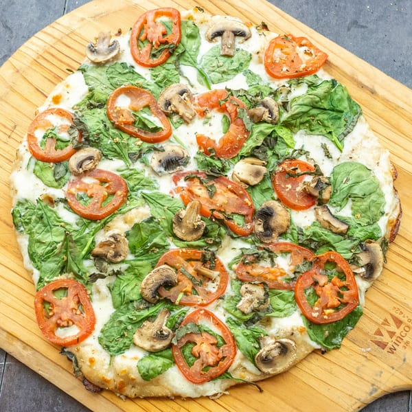 Mama Mimi's Take 'N Bake Pizza with spinach, tomatoes, and mushrooms on a wooden board.