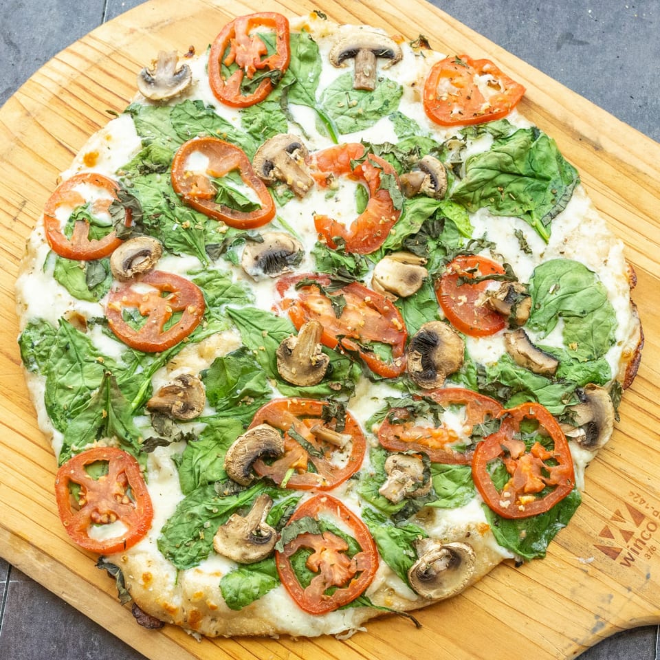 Mama Mimi's Take 'N Bake Pizza with spinach, tomatoes, and mushrooms on a wooden board.