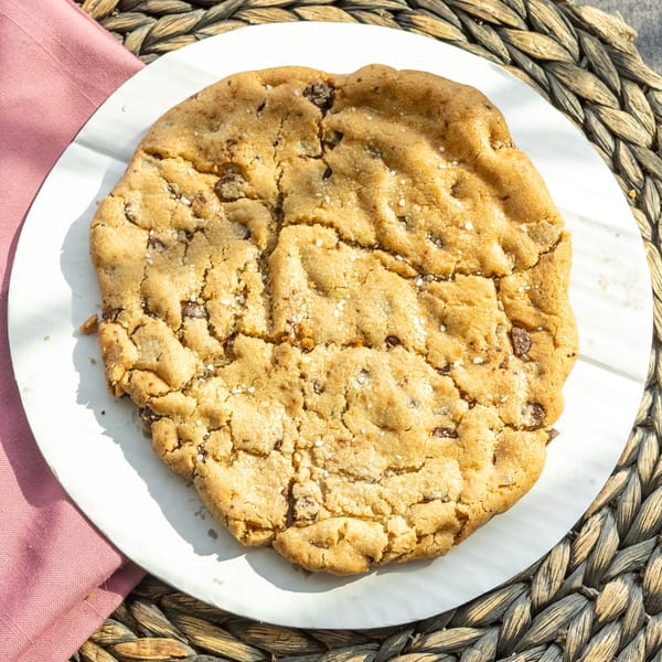 A large, golden-brown chocolate chip cookie with sea salt on a white plate and woven placemat.