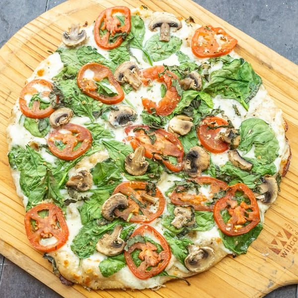 Mama Mimi's Take 'N Bake Pizza with spinach, sliced tomatoes, and mushrooms on a wooden peel.